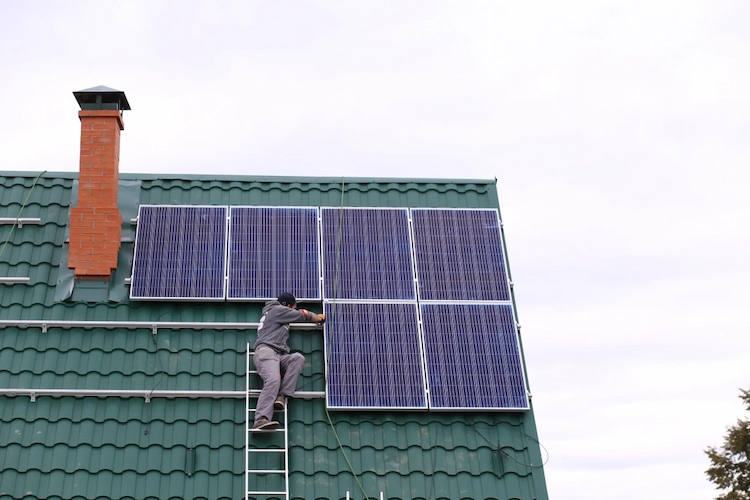 installer of solar panels works on sloping roof with ladder