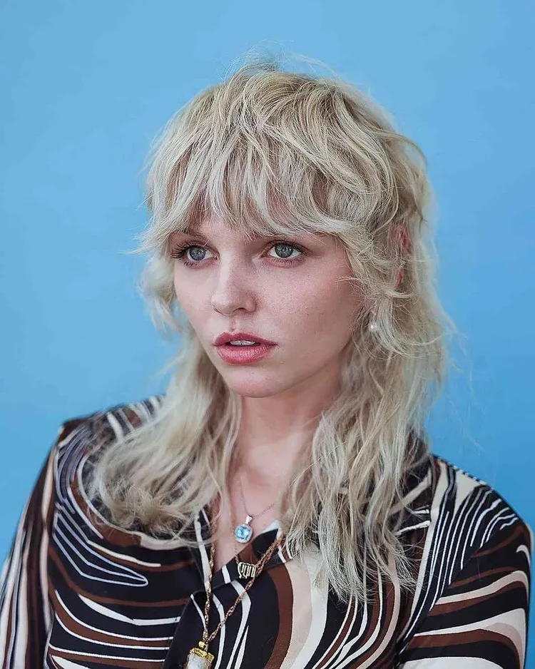 The blonde shullet hairstyle with bangs is inspired by the 70s era